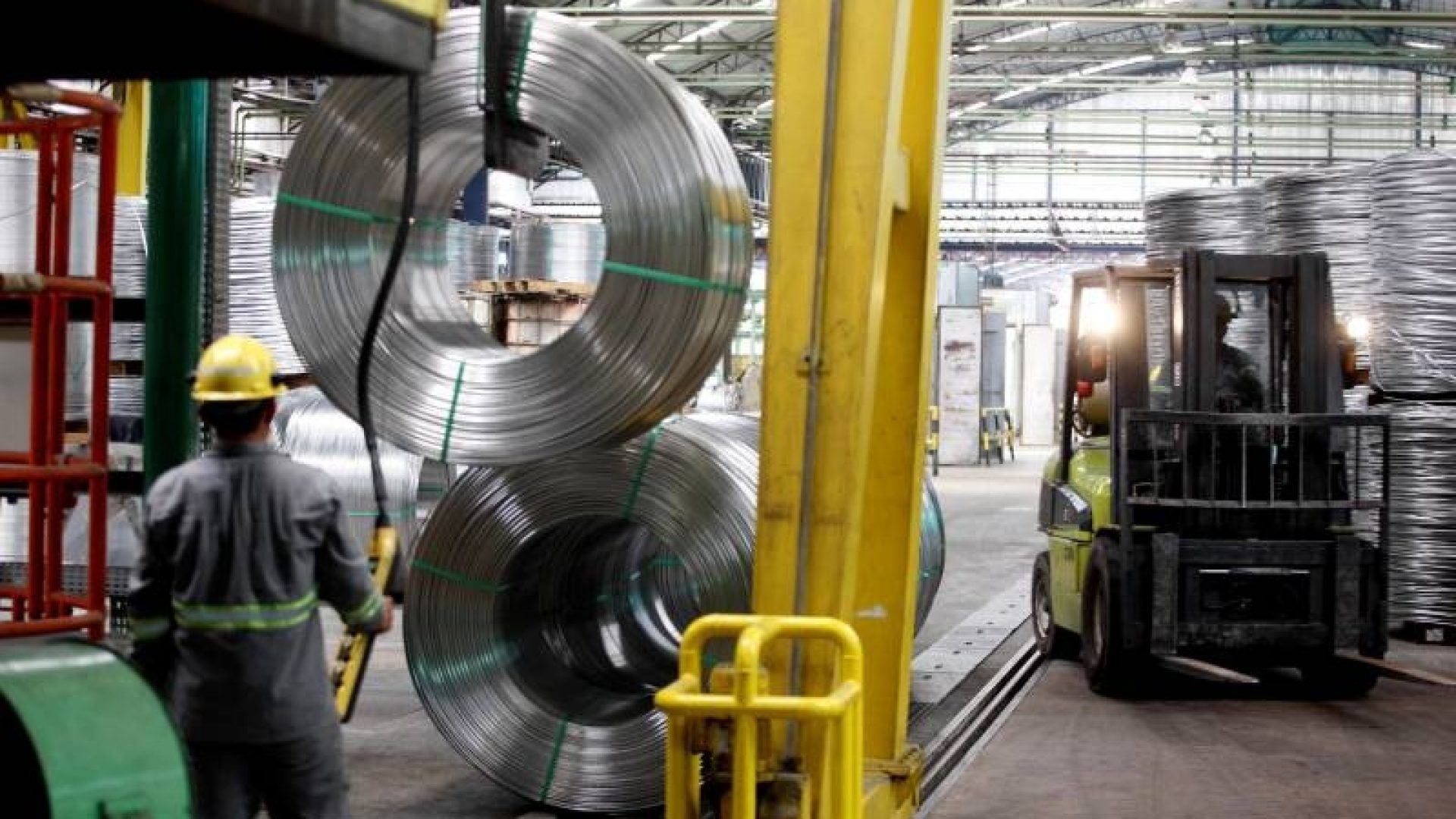 Trabalhador com capacete amarelo manuseando grandes bobinas de metal em um galpão industrial.