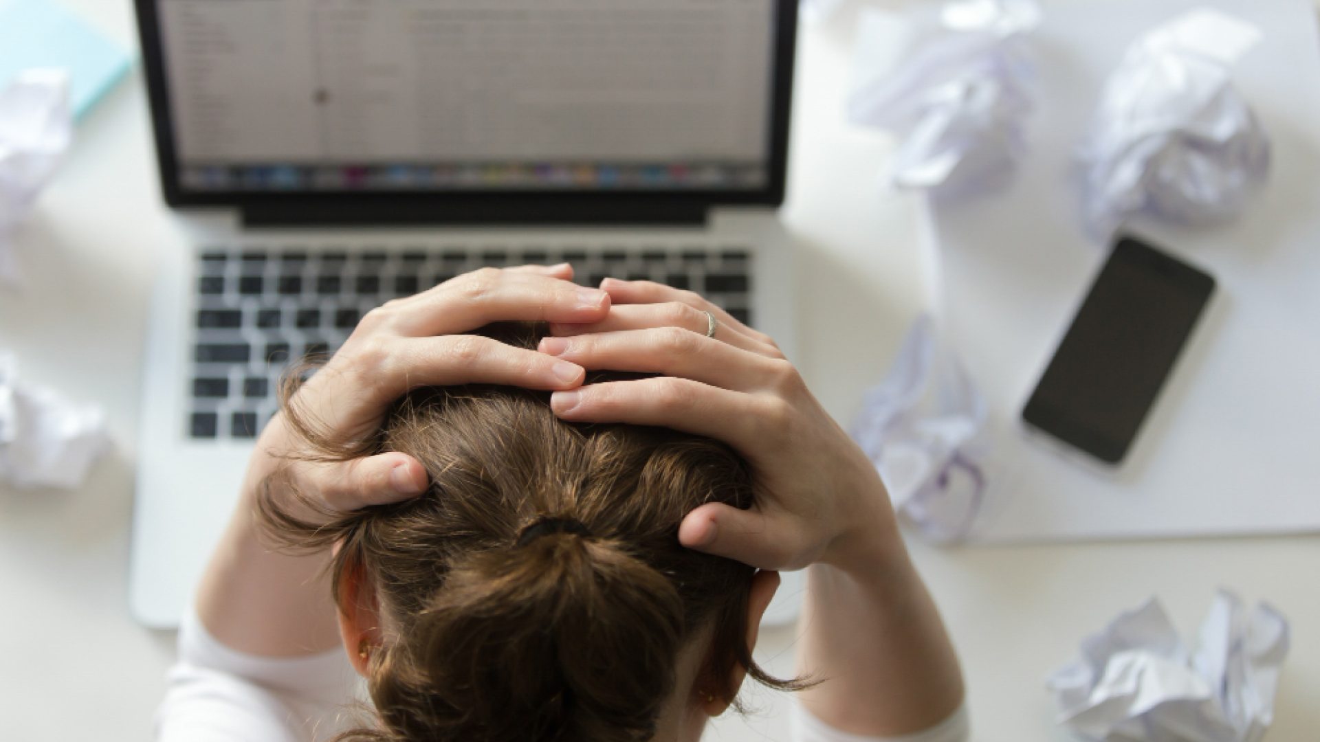Pessoa com as mãos na cabeça em frente a um notebook, com papéis amassados.