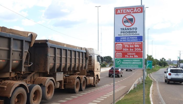 Um caminhão caçamba sujo passando por uma placa que proíbe o trânsito de veículos pesados em horários específicos.