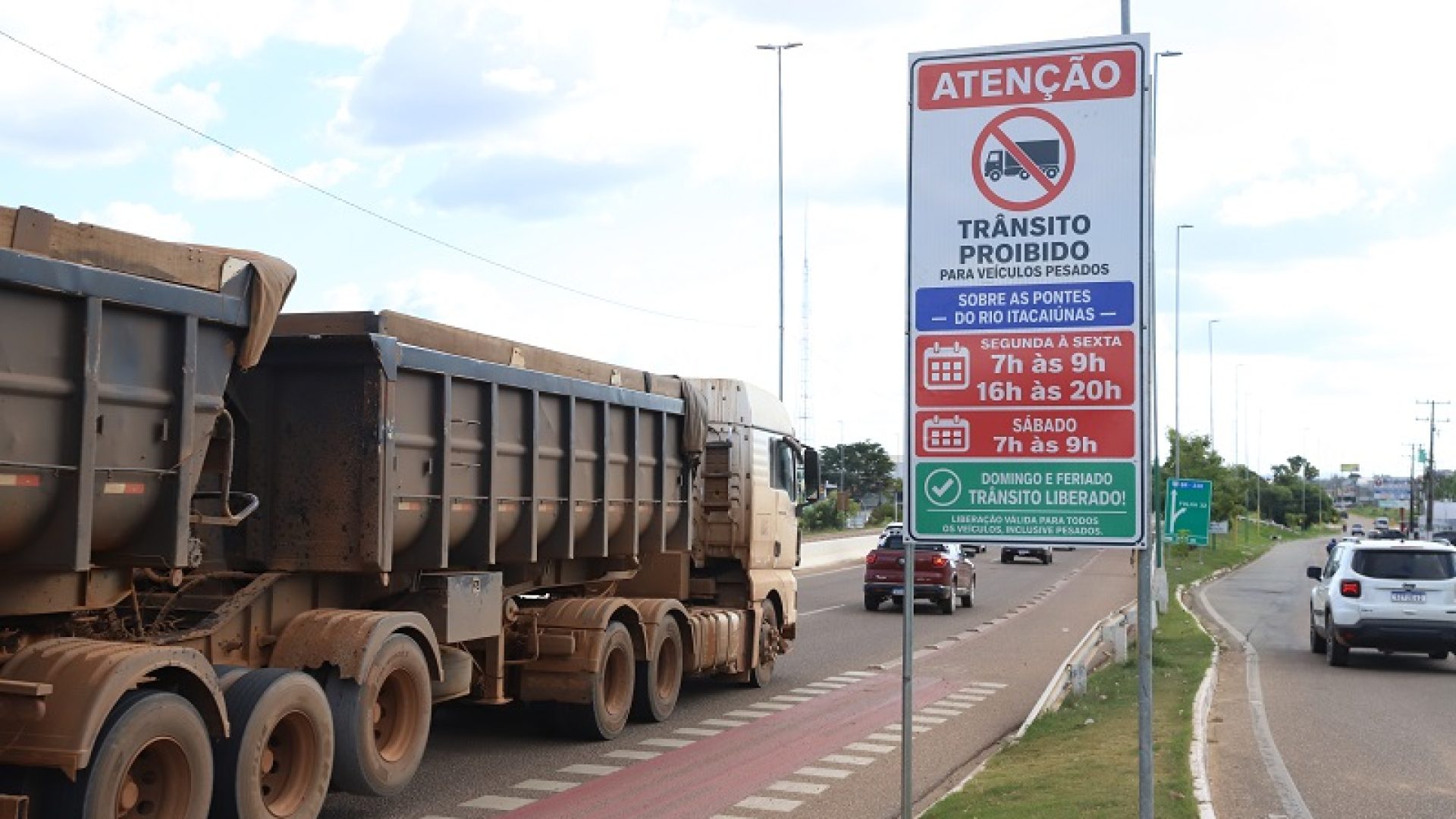 Um caminhão caçamba sujo passando por uma placa que proíbe o trânsito de veículos pesados em horários específicos.