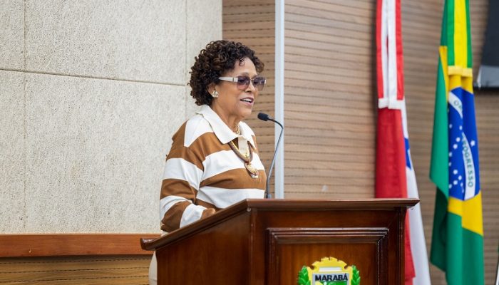 Mulher negra de cabelos cacheados curtos, vestindo blusa listrada, discursando em um púlpito de madeira com o brasão de Marabá.